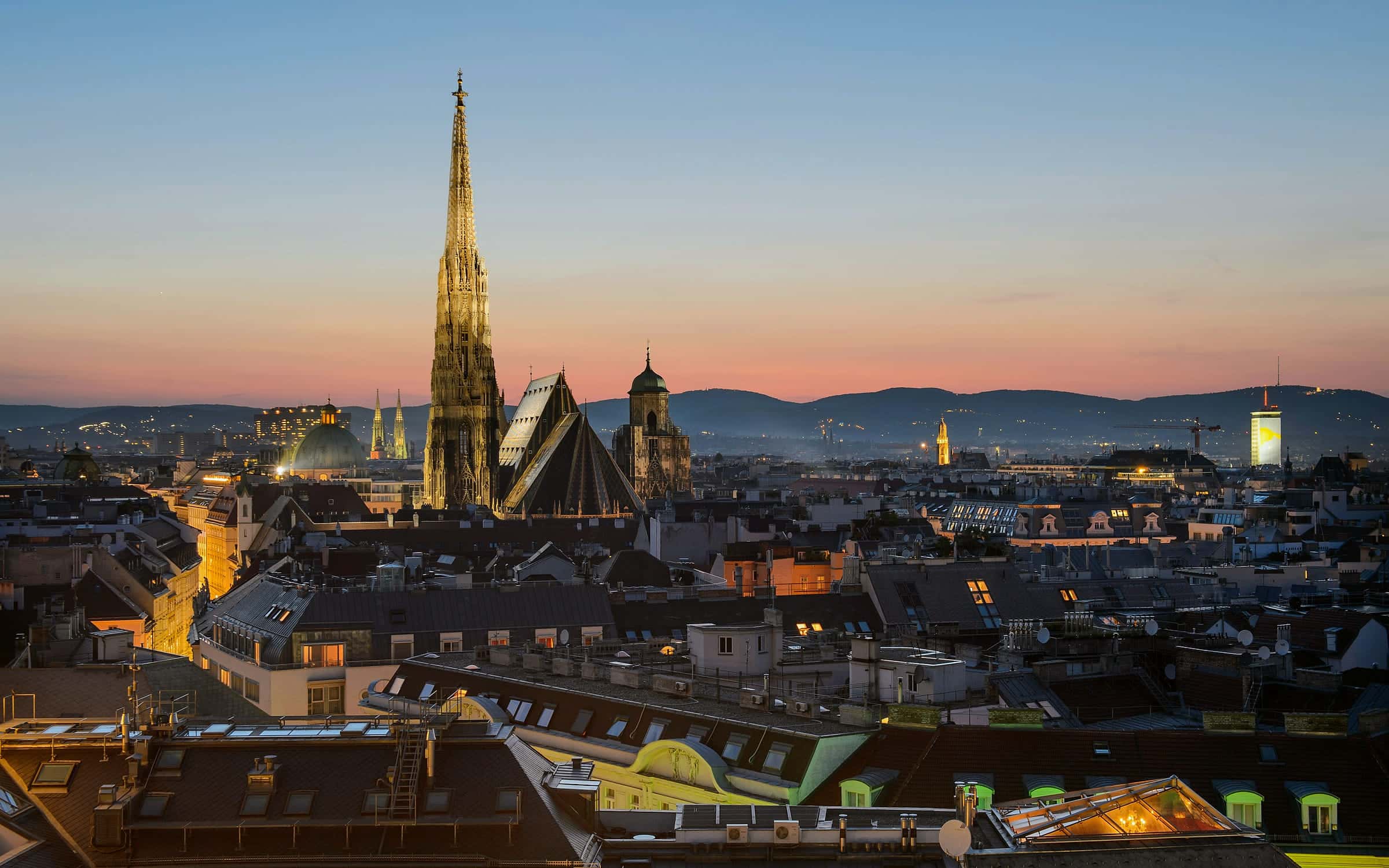 Die Dächer der Wiener Innenstadt im Abendlicht, dahinter der beleuchtete Steffl und der Horizont über dem Wienerwald.